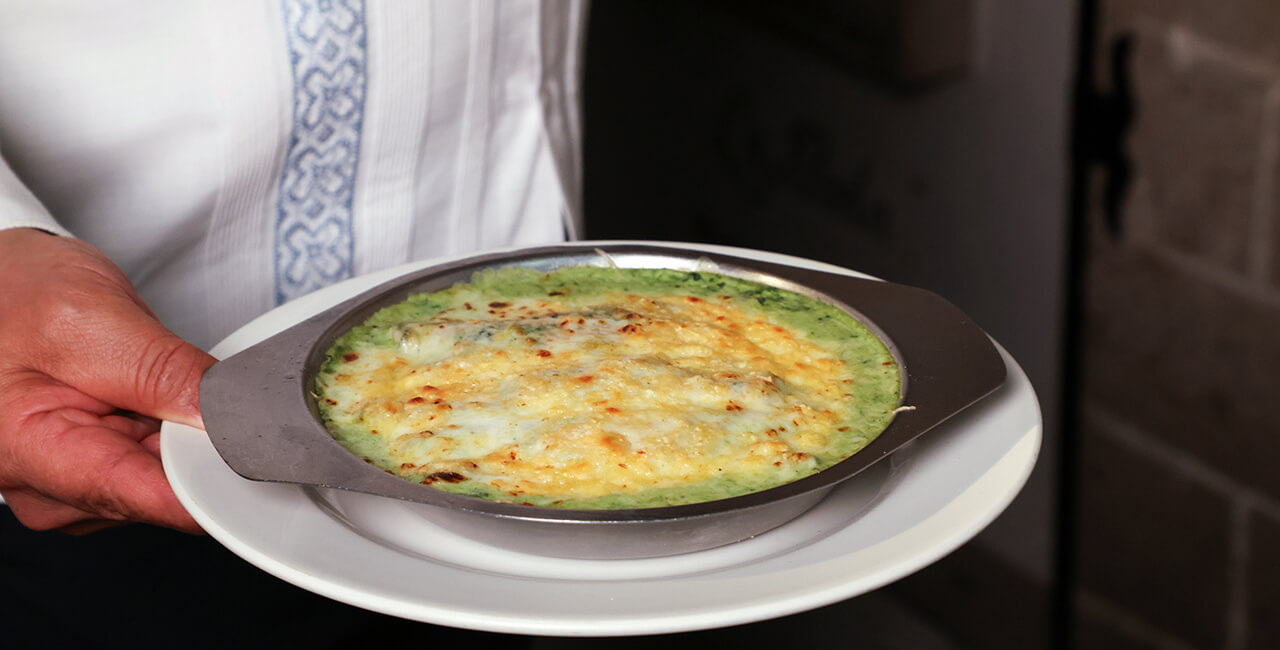 Mesero con camisa t&iacute;pica del uniforme del Caf&eacute; de Tacuba sirviendo un plato de enchiladas verdes gratinadas.