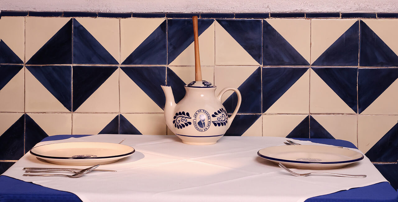 Mesa servida en el Caf&eacute; de Tacuba con manteler&iacute;a y vajilla con el logotipo del restaurante, frente a mosaicos t&iacute;picos de la pared, sin alimentos.