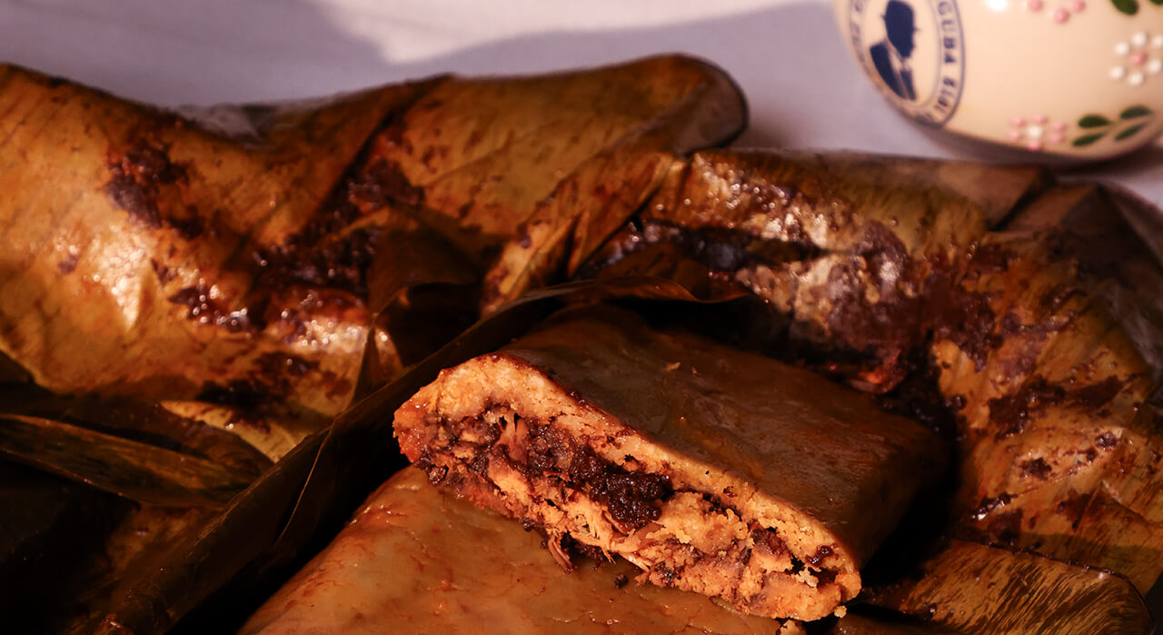 Tamal oaxaque&ntilde;o abierto en hoja de pl&aacute;tano, relleno de pollo con mole, con una taza con el logotipo del Caf&eacute; de Tacuba al fondo.