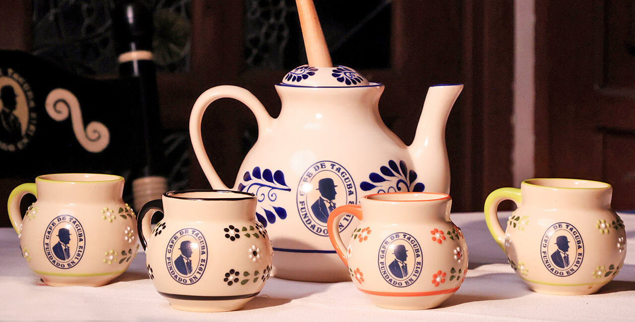 Jarra y cuatro tazas con el logotipo del Caf&eacute; de Tacuba sobre una mesa del restaurante, con molinillo de madera para espumar chocolate.