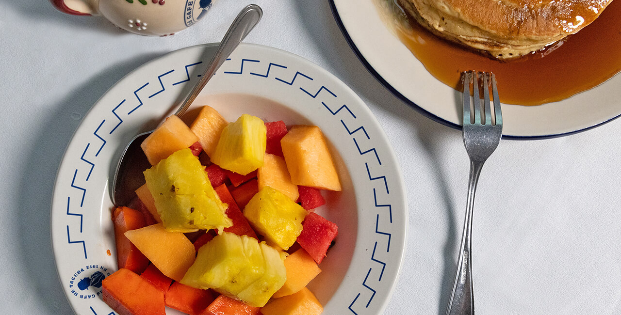 Plato del men&uacute; infantil con fruta picada y hot cakes con miel, en toma cenital, con cubiertos a punto de comer en el Caf&eacute; de Tacuba.